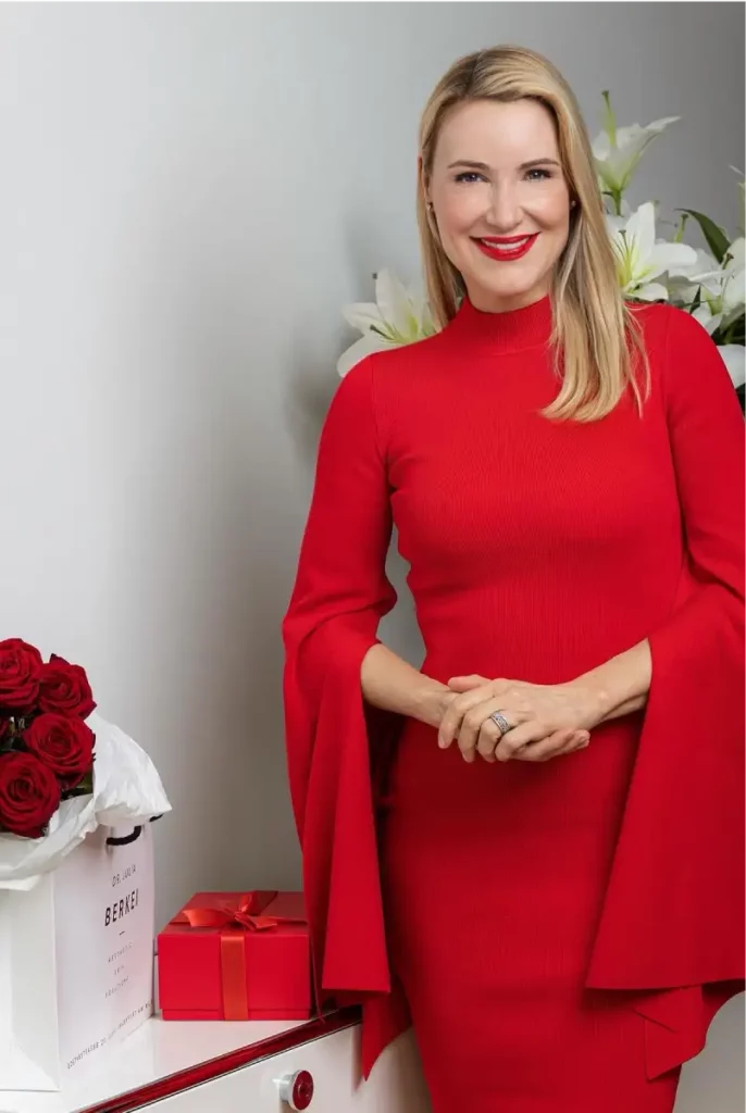 Aufnahme von glamouröser Dr. Berkei im roten Kleid neben einem Strauß roter Rosen und einem roten Geschenk.