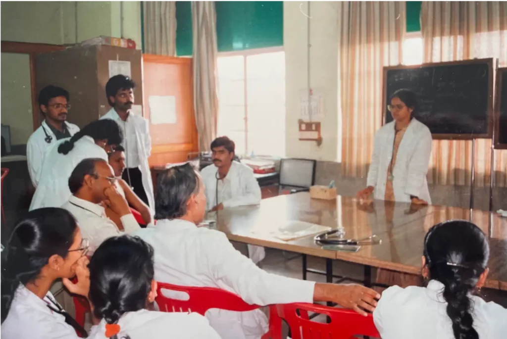 Aufnahme von einer Art Klassenzimmer. Eine Person steht vorne vor einer Tafel und alle anderen sitzen und stehen weiter hinten. Alle tragen einen weißen Kittel und ein Stetoskop um den Hals.