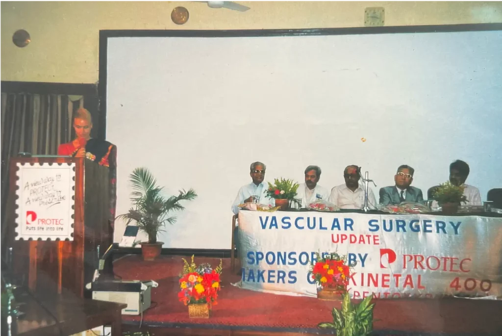 Dr. Berkei, die einen Vortrag an einem Podium hält. Rechts von sitzen fünf Männer an einem Tisch mit einem Banner "VASCULAR SUGERY BANNER".